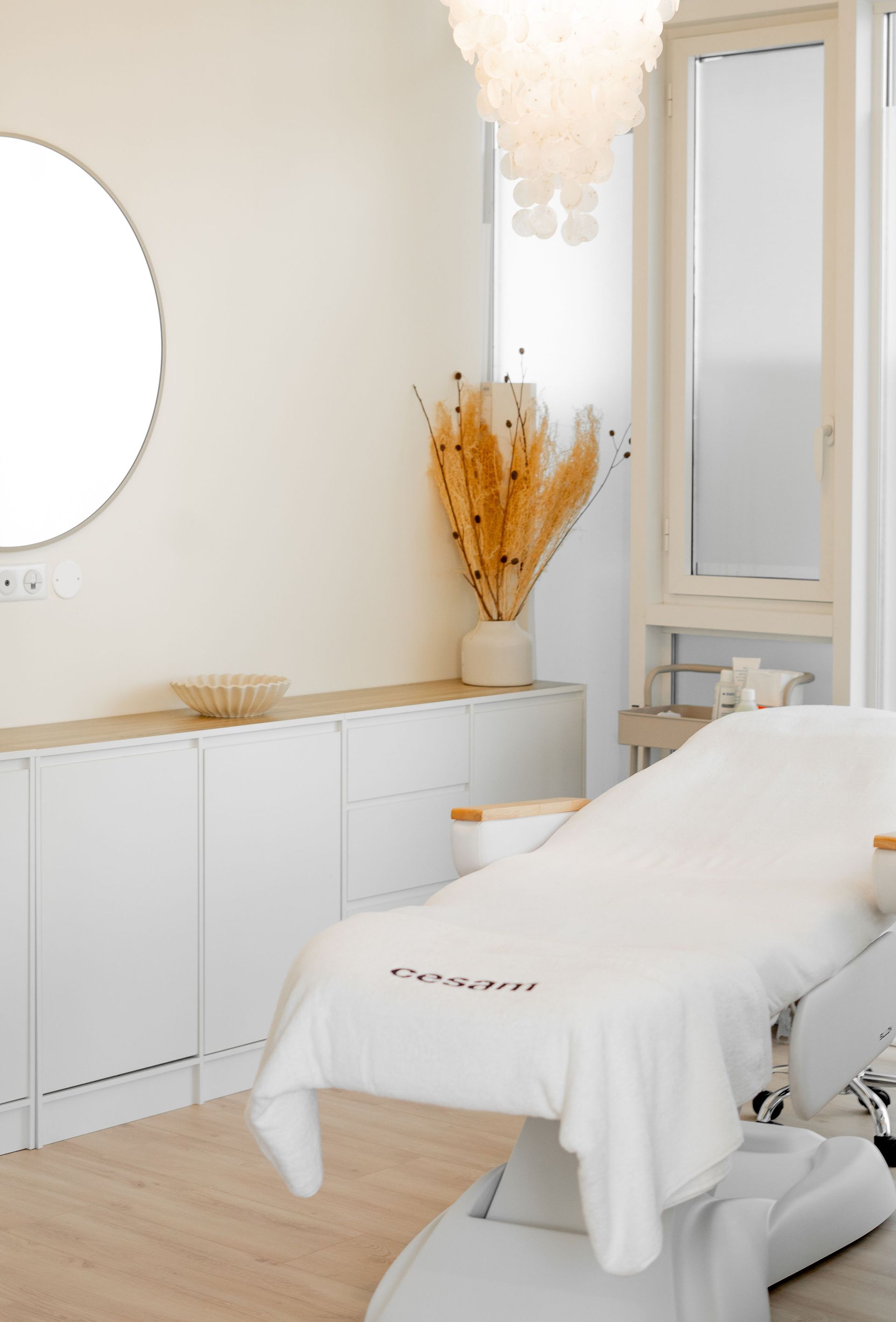 Salle de spa lumineuse avec une table de soin blanche, des placards, un miroir et une décoration florale séchée