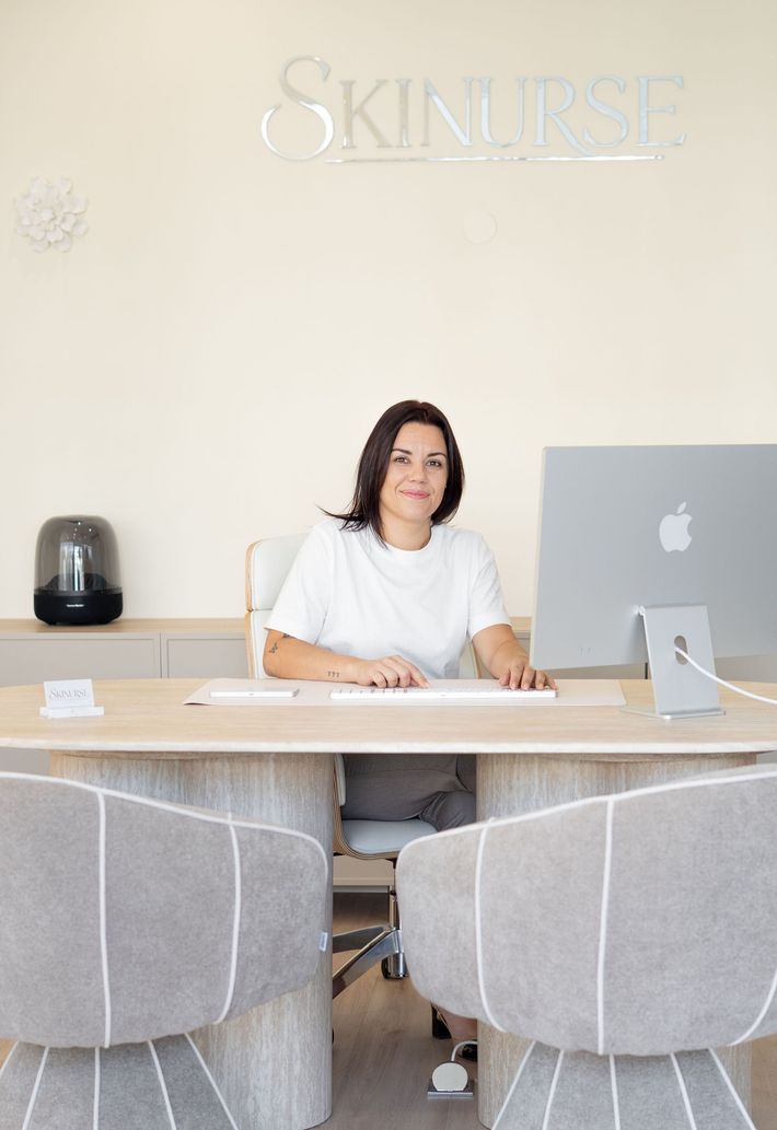 Femme assise à un comptoir d’accueil dans le hall lumineux d’un salon à la décoration neutre, avec deux fauteuils gris