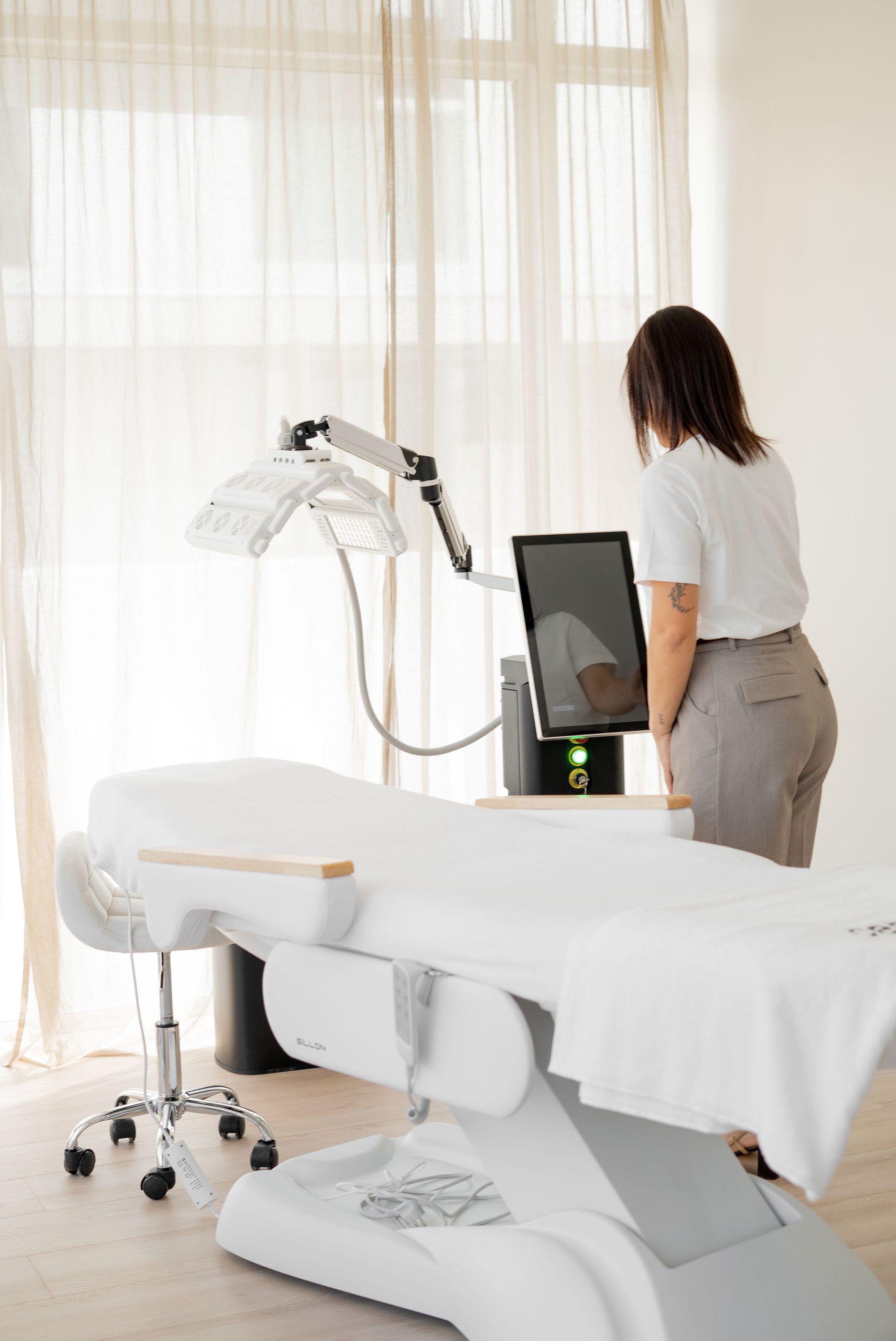 Femme debout à côté d’une table de soin dans une salle de spa lumineuse, en train de régler un appareil de soin du visage