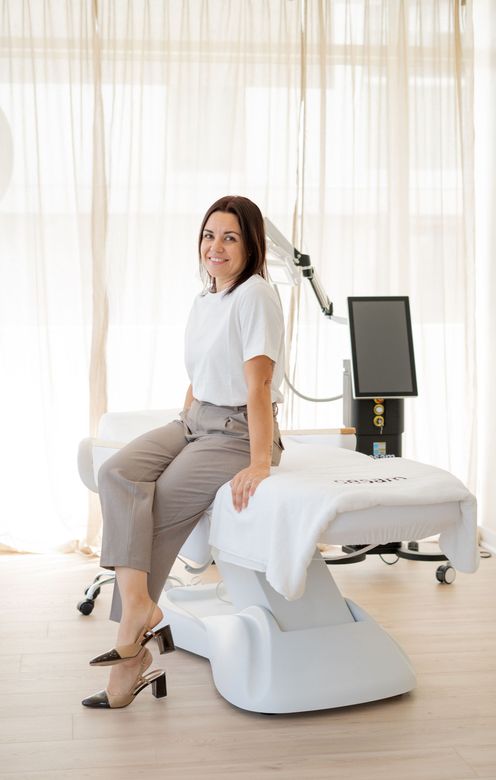 Une femme assise sur une table de soins blanche dans une salle de clinique lumineuse, souriant à la caméra.