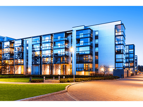Edificio de apartamentos moderno de cristal al anochecer, con ventanas iluminadas y una carretera curva enfrente.