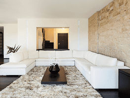 Sala de estar moderna blanca con sofá modular, alfombra de pelo largo y mesa de centro oscura frente a una pared decorativa de piedra.