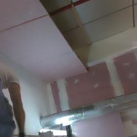 Una persona de pie en una habitación con el techo dañado y las paredes rosas cerca de un objeto metálico.