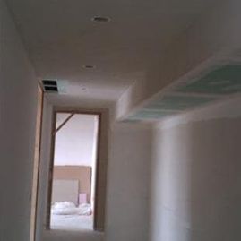 Pasillo estrecho sin terminar, con paredes blancas, rejillas de ventilación en el techo y una puerta que da a una habitación con materiales a la vista.