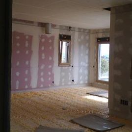 Habitación parcialmente renovada con parches de yeso rosa, suelo a la vista y ventanas que dejan entrar la luz natural.