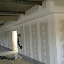 Habitación interior en construcción con paneles de yeso, techo a la vista y un trabajador en una abertura de la pared.