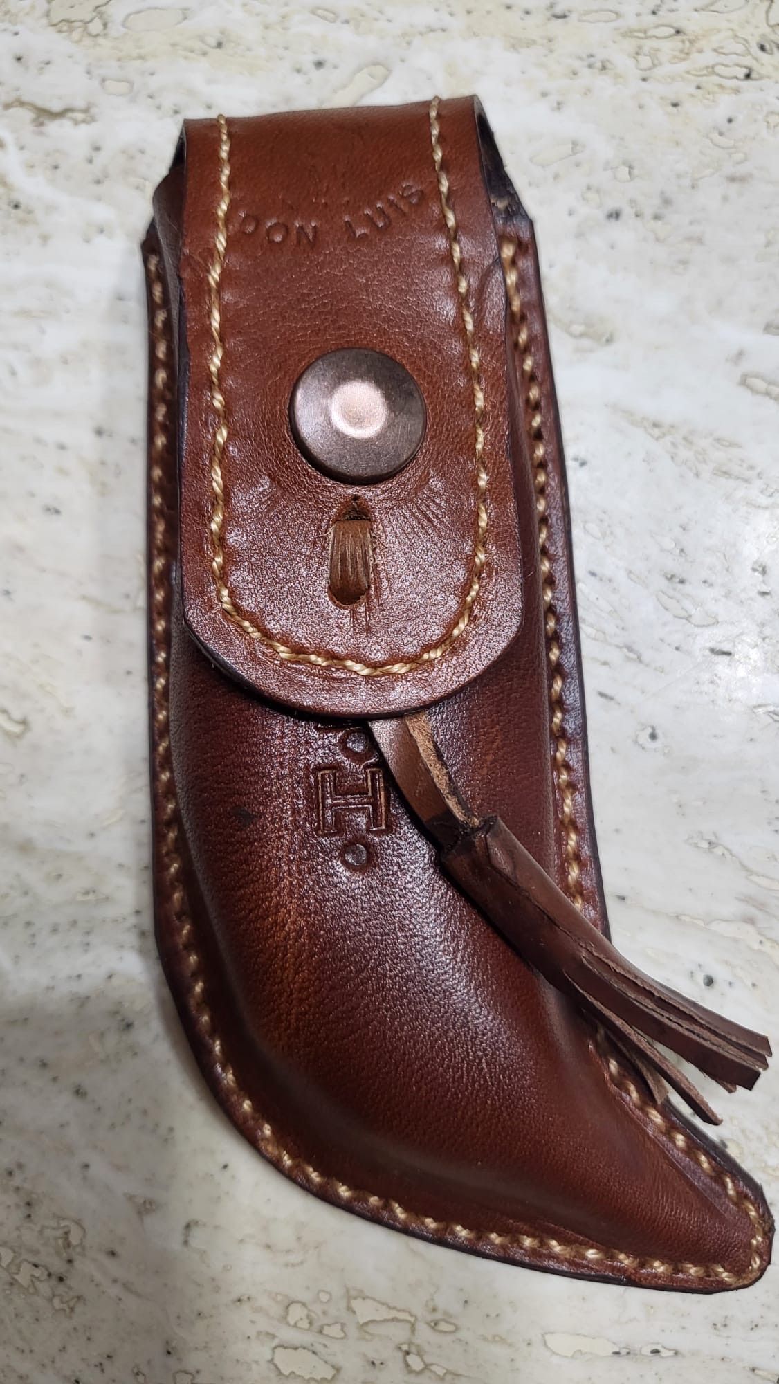 Funda de cuero marrón con solapa de cierre a presión y borla, colocada en vertical sobre una superficie clara
