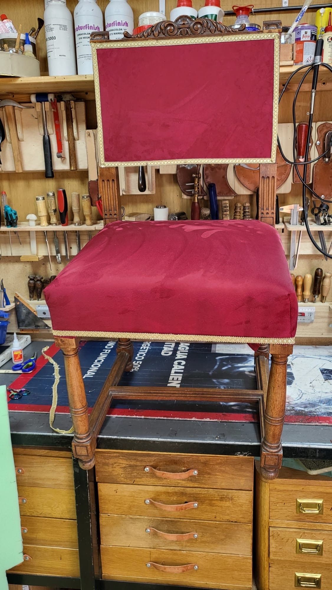 Silla tapizada de rojo en un taller de carpintería, con herramientas y estantes al fondo.