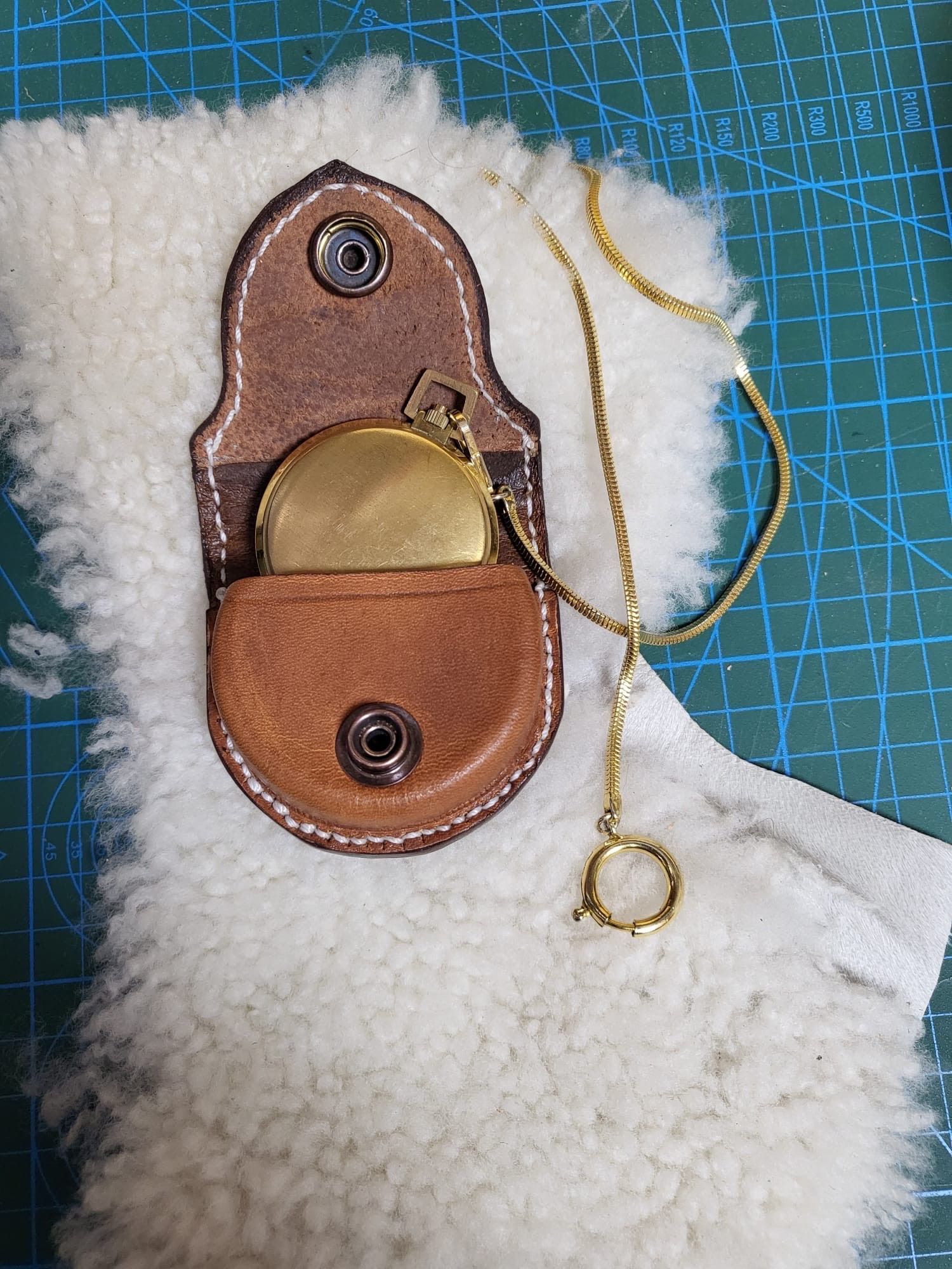 Bolsa de cuero marrón con cierre a presión de latón sobre una prenda de tejido polar blanco, con un cordón dorado y una anilla redonda para tirar.