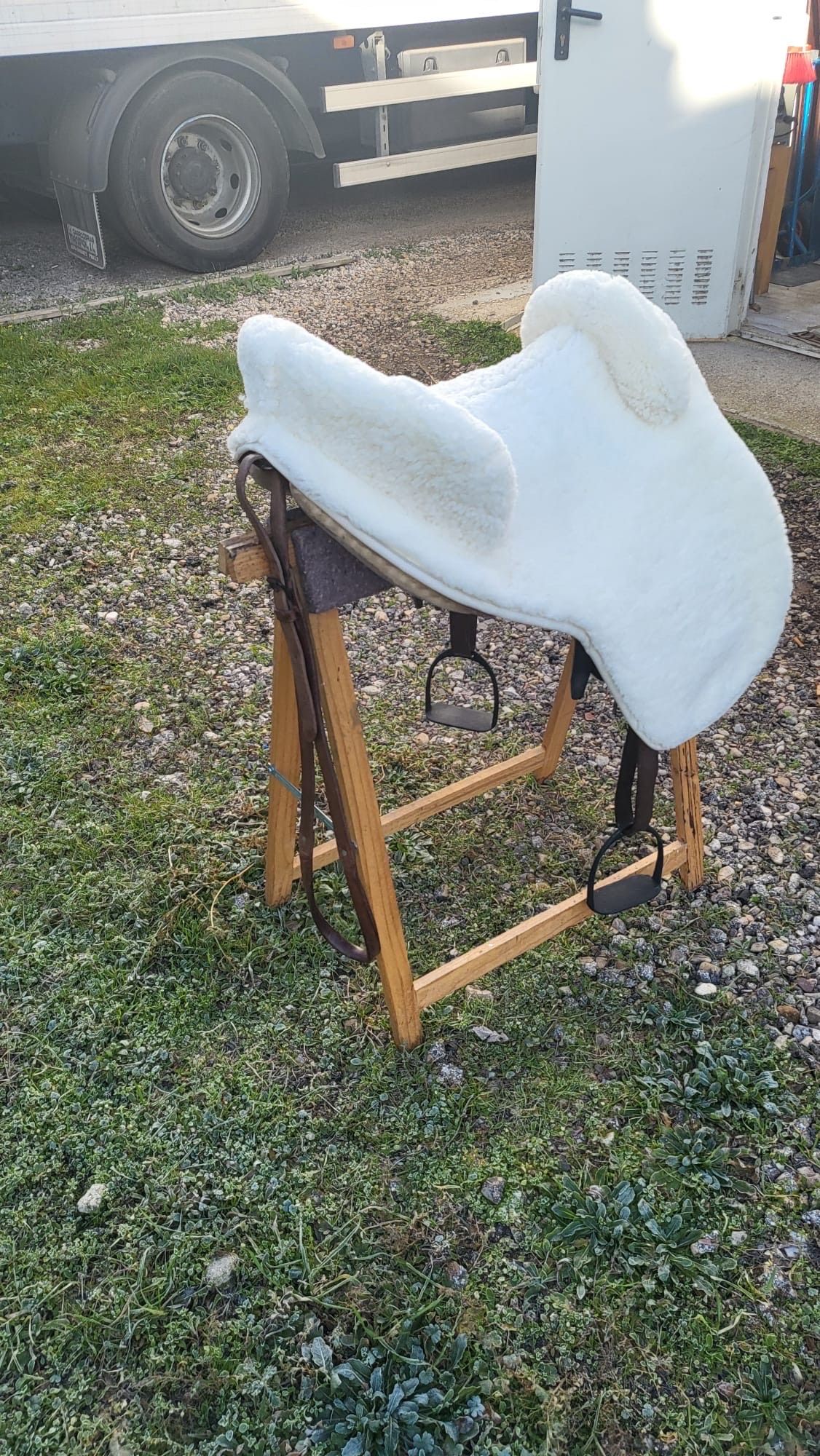 Una mantilla blanca sobre un soporte de madera para sillas de montar, al aire libre sobre la hierba, junto a un remolque utilitario.