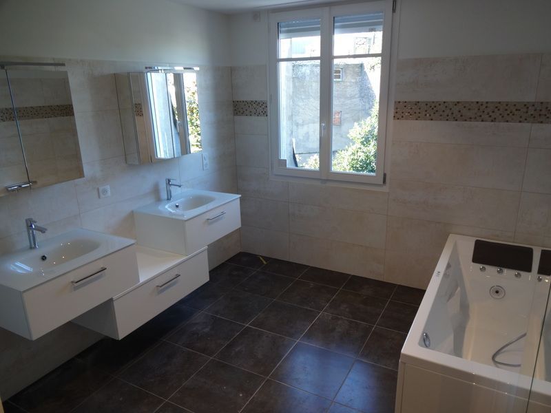 Salle de bains avec double vasque, grand miroir, fenêtre et baignoire sur sol carrelé.
