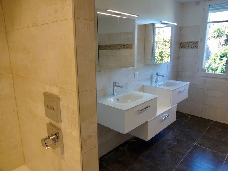 Salle de bains moderne avec deux lavabos blancs, de grands miroirs, des murs carrelés beiges et un carrelage foncé au sol.