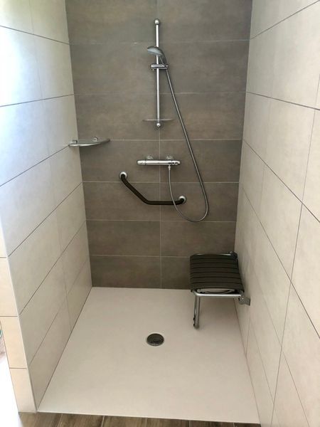 Douche accessible avec barres d'appui, pommeau de douche à main, siège intégré et murs carrelés.