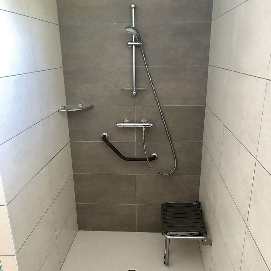 Cabine de douche accessible avec barre d'appui, douchette et petit siège sur murs carrelés