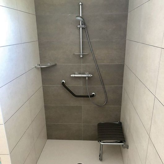 Cabine de douche accessible avec barre d'appui, douchette et petit siège sur murs carrelés
