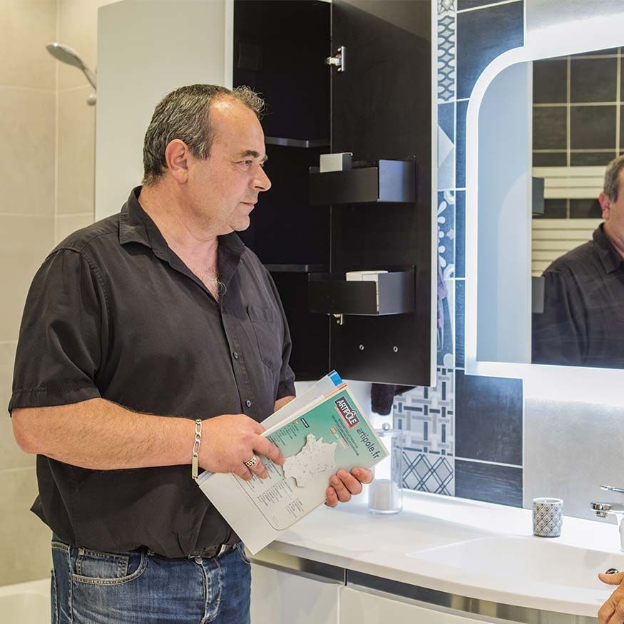 Un homme tenant un livret à côté d'un lavabo et d'un miroir de salle de bains, regarde les étagères de la coiffeuse.