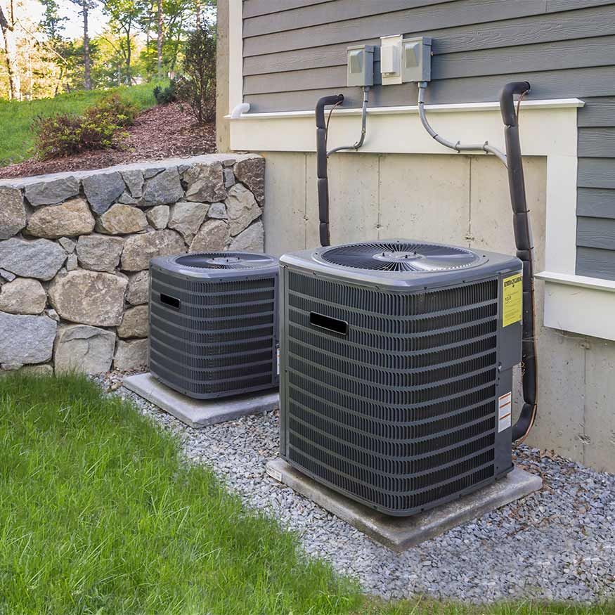 Deux unités de climatisation extérieures à côté d'une maison sur un terrain gravillonné, avec des lignes électriques et un mur de soutènement en pierre.
