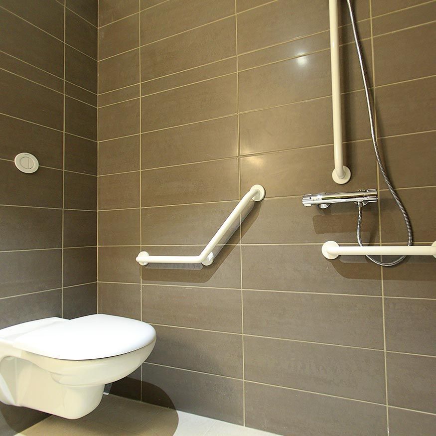 Salle de bain accessible avec murs carrelés marron, lavabo suspendu et barres d'appui blanches autour de la douche.
