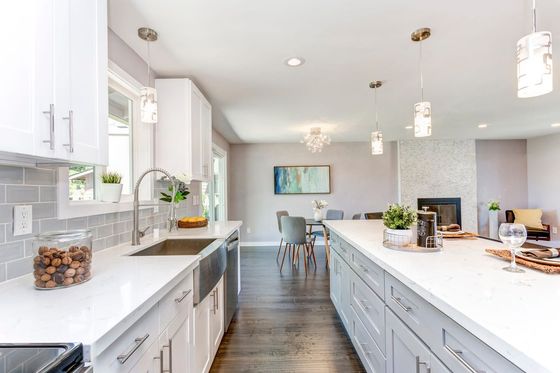 Cocina moderna y luminosa con gabinetes blancos, encimeras de cuarzo y una gran isla. Pisos de madera oscura y una chimenea al fondo.