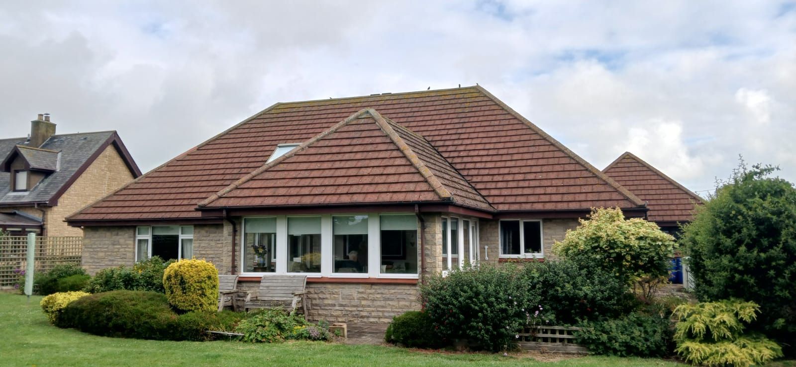 A brick house with a brown tiled roof, windows, and bushes in front. A cloudy sky is overhead.
