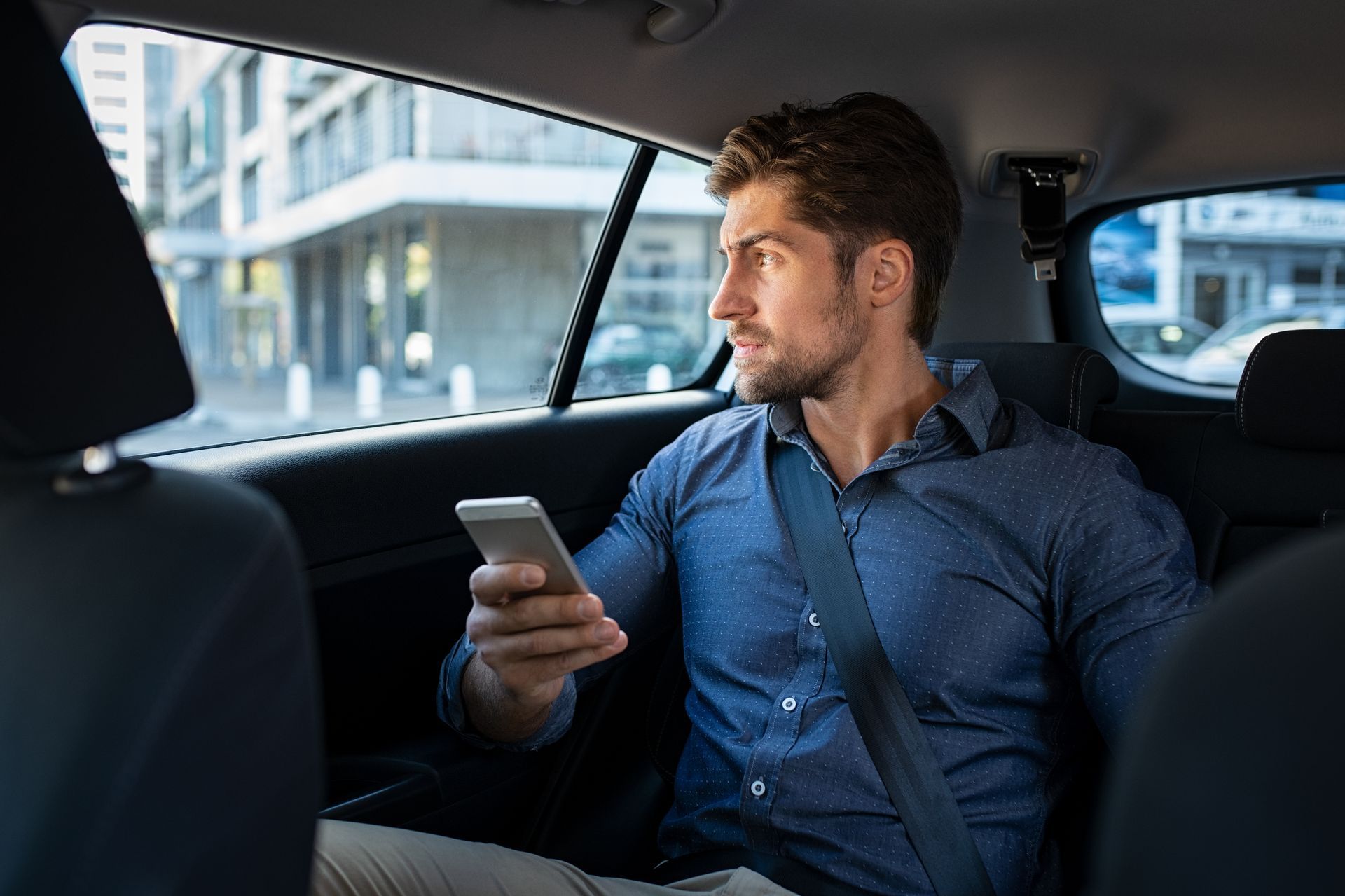 Un homme en chemise bleue, assis dans une voiture, regarde par la fenêtre tout en tenant un smartphone.