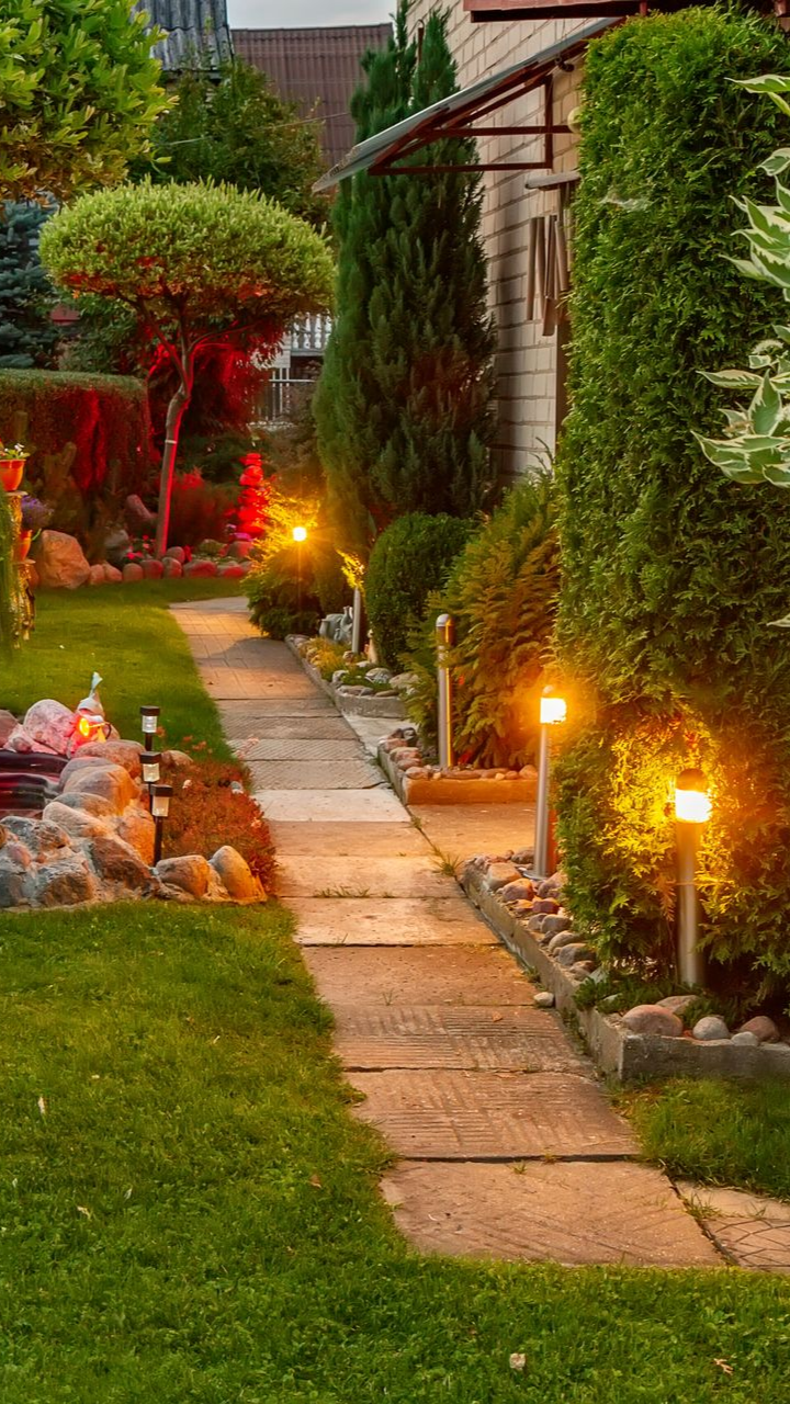 Chemin en pierre éclairé par des lumières chaudes, flanqué d'herbe verte, de buissons et d'arbres.