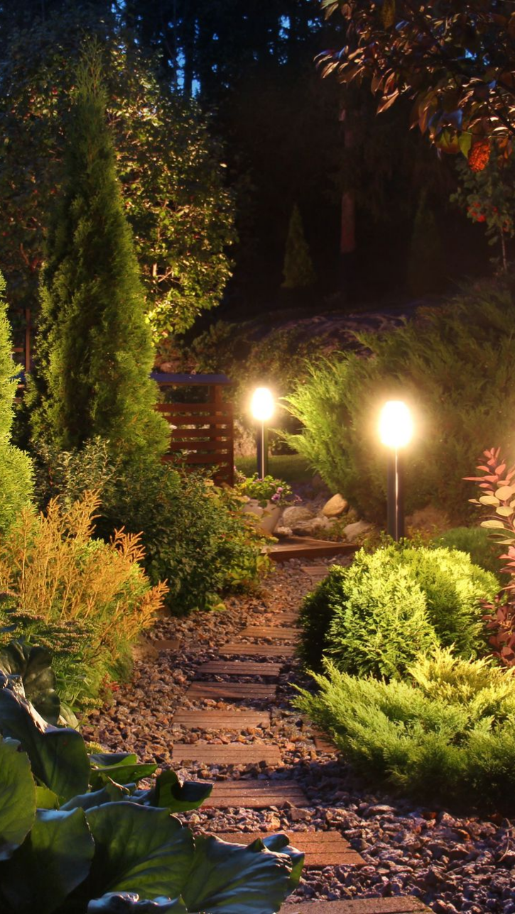 Un chemin de pierre éclairé par des orbes lumineux traverse un jardin luxuriant la nuit.