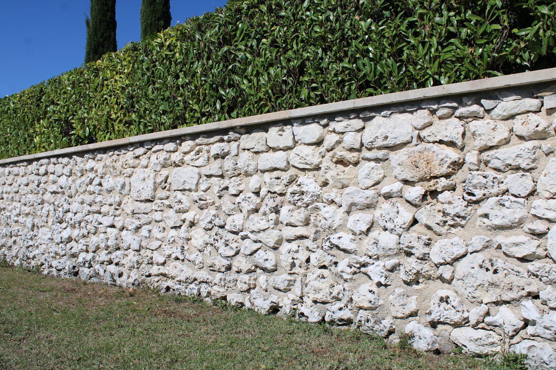 Mur de pierre surmonté d'une haie verte contre un ciel bleu.