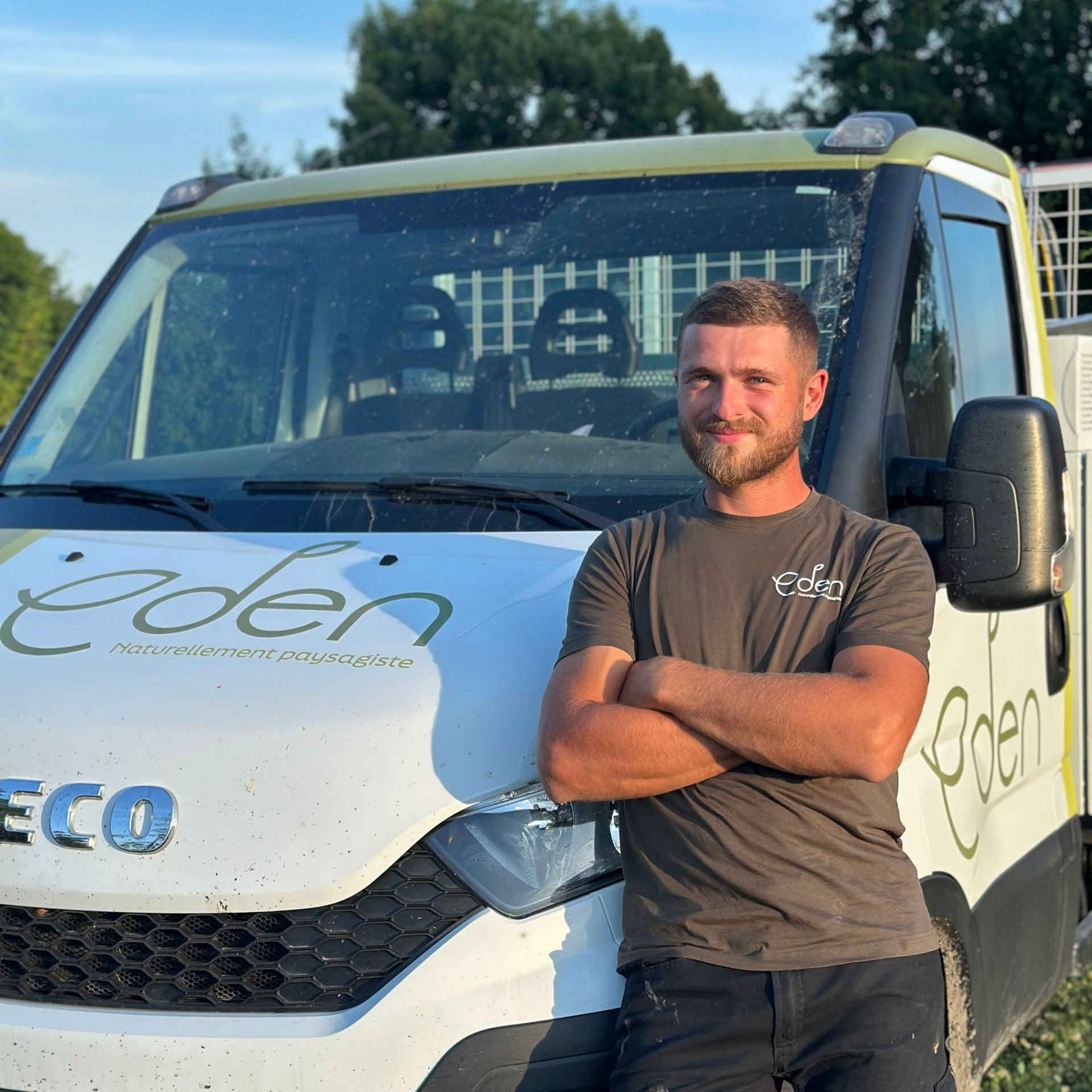 Un homme aux bras croisés, souriant, debout près d'une camionnette de travail blanche Eden Paysage.