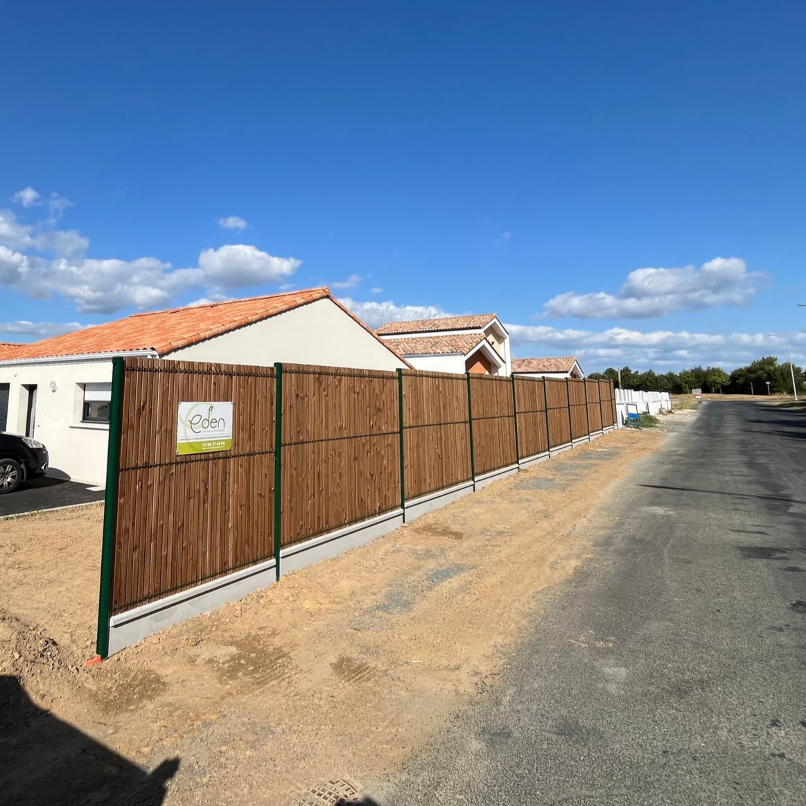 Clôture en bois le long d'une route devant une maison sous un ciel bleu.