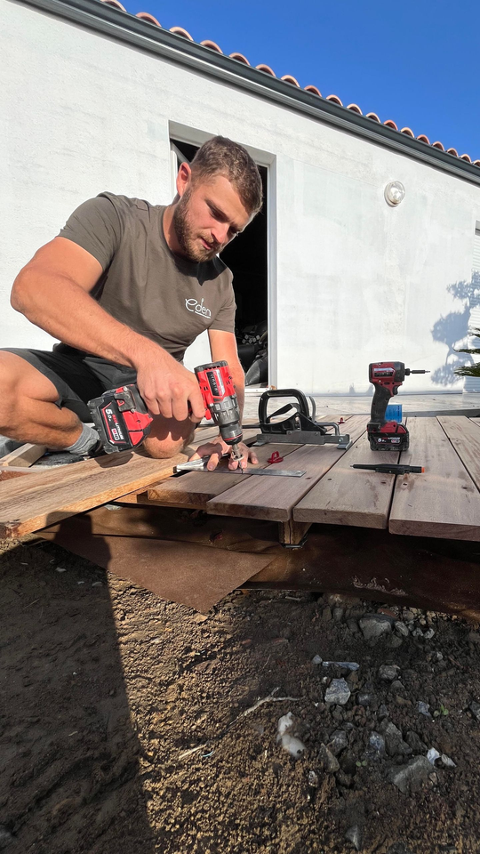 Un homme utilise une perceuse pour travailler sur une terrasse en bois à l'extérieur ; journée ensoleillée.