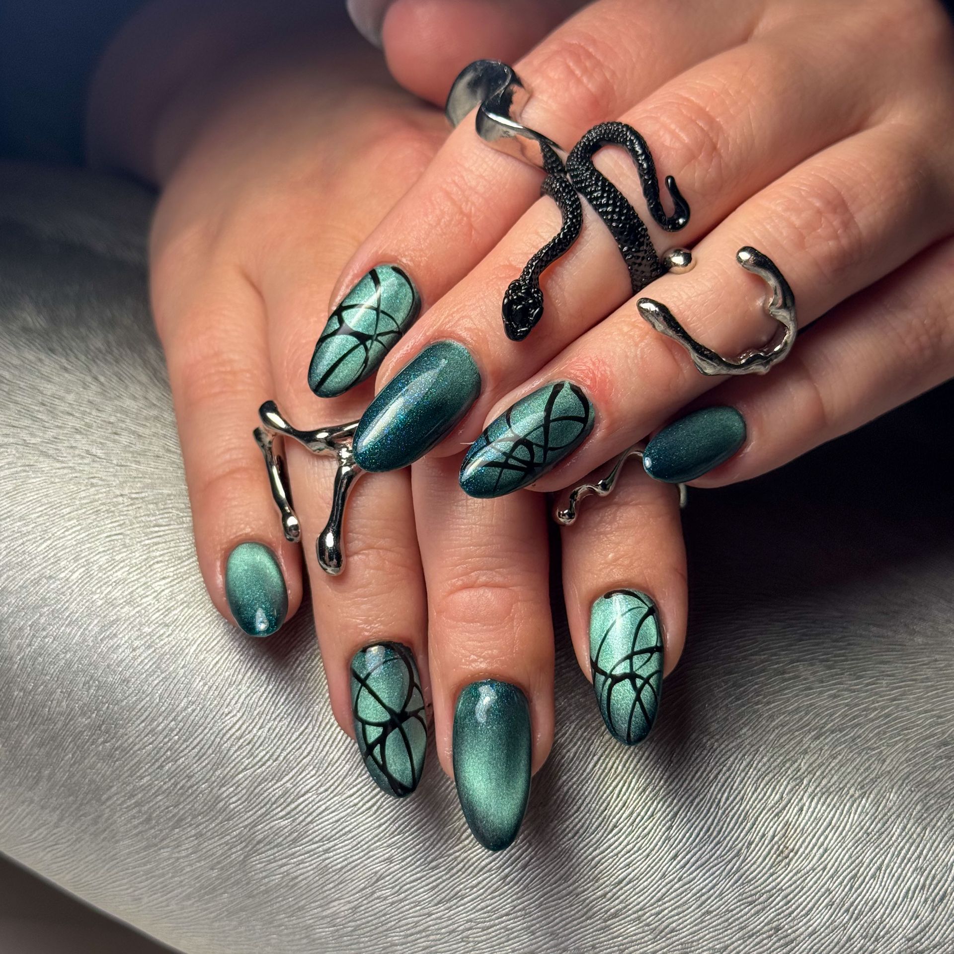 Hand mit lackierten Nägeln in Grün-, Gold- und Brauntönen, in der Nähe von getrockneten Blumen.