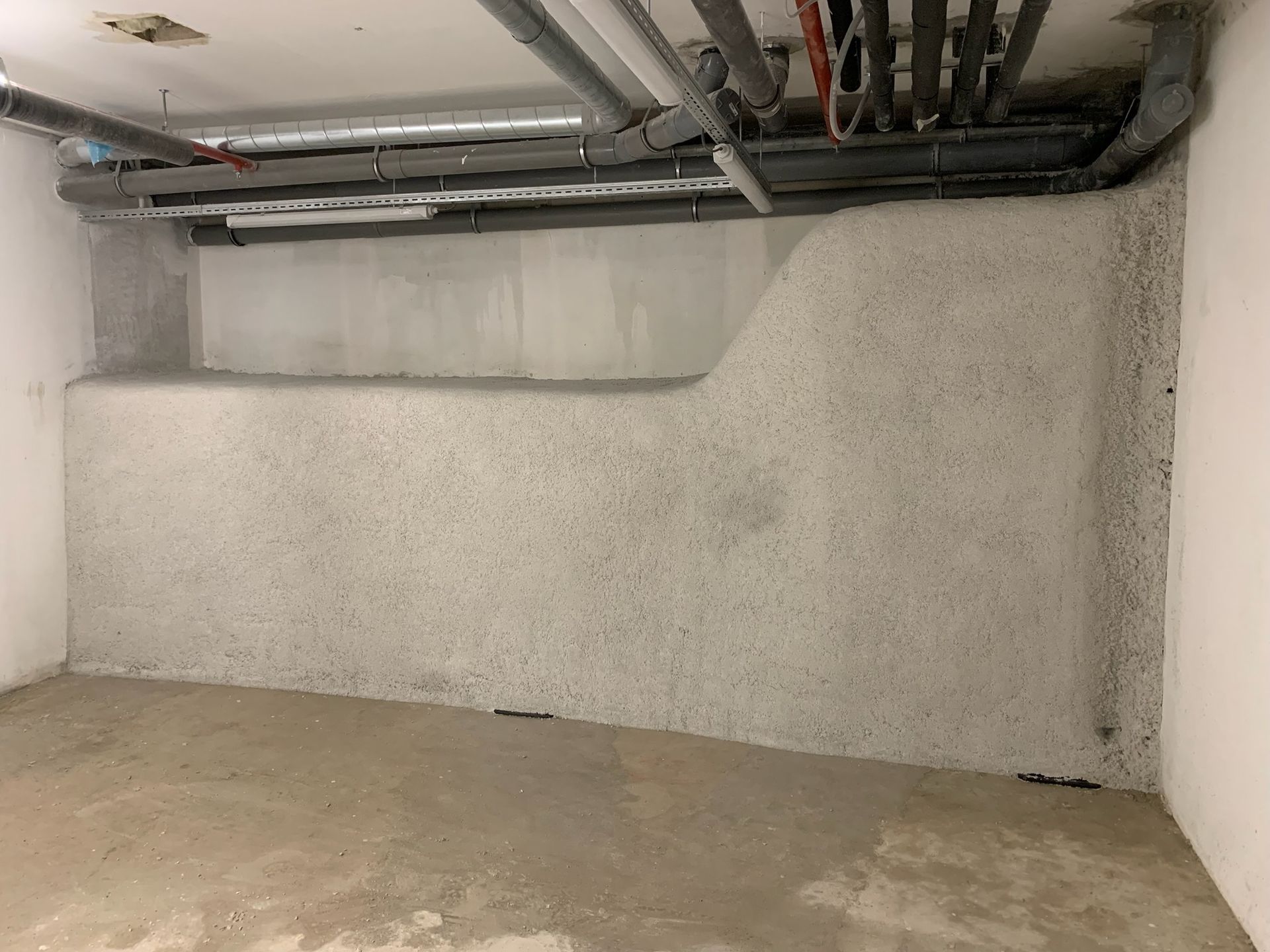 Mur de ciment gris avec une partie surélevée, tuyaux au-dessus, dans une pièce inachevée.