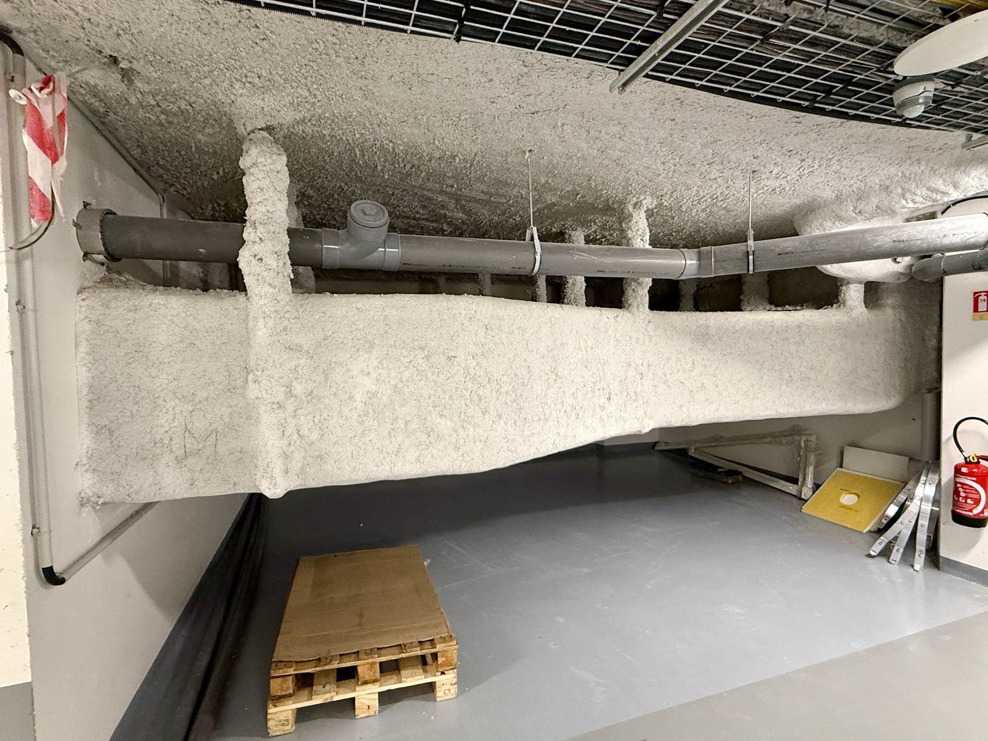 Une poutre en béton et des tuyaux recouverts d'un produit ignifuge blanc projeté dans un sous-sol. Une palette en bois est posée au sol.
