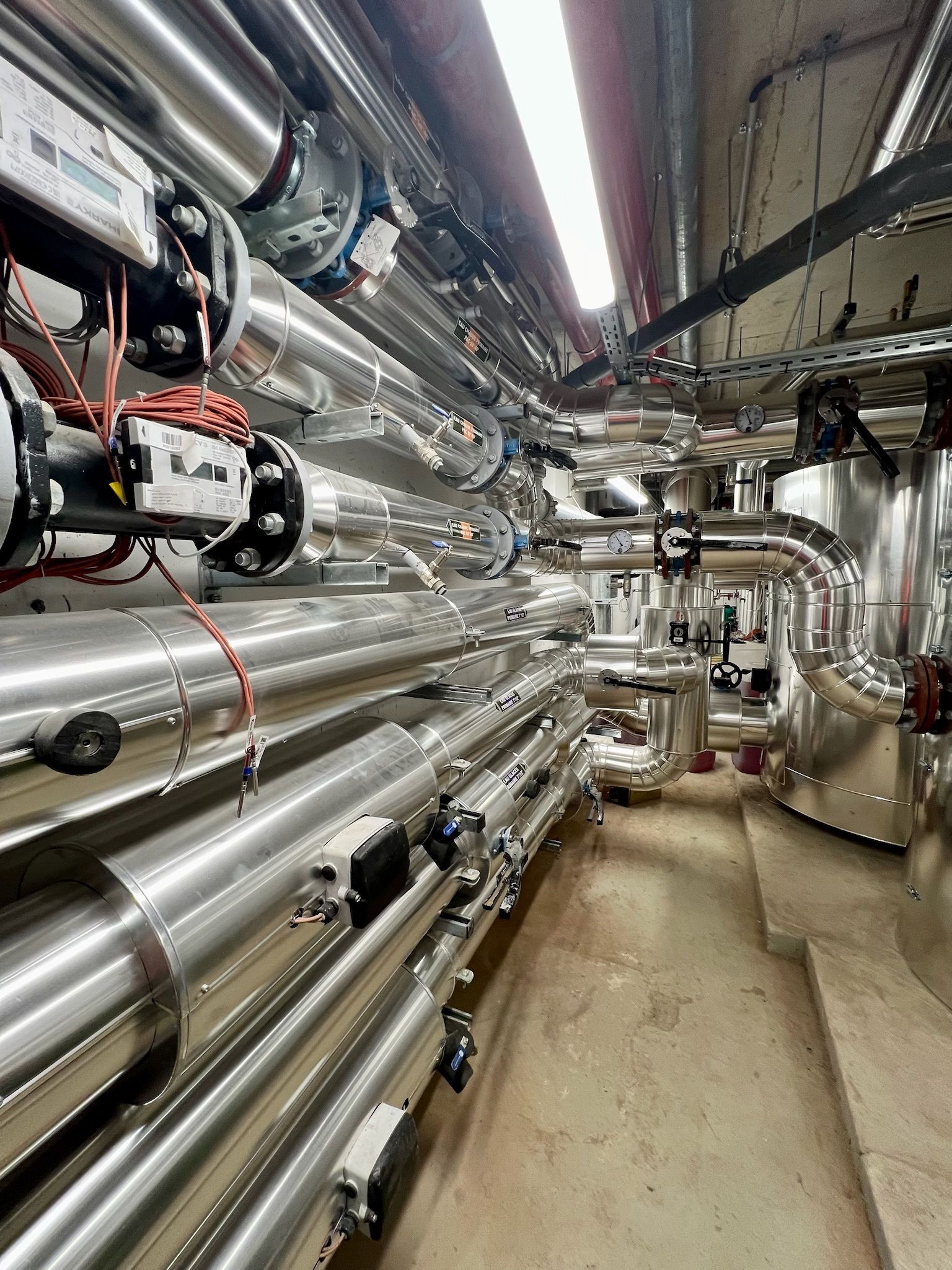 Des rangées de tuyaux métalliques brillants dans une buanderie, avec du câblage et d'autres équipements, éclairés par des plafonniers.