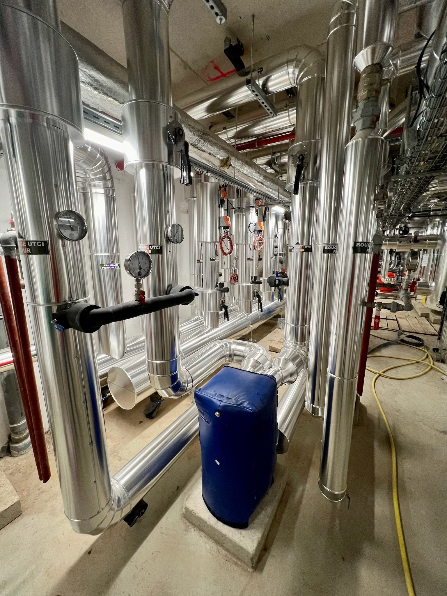 Tuyauteries et équipements mécaniques dans un sous-sol ou une buanderie. Tuyauteries argentées, équipements bleus et murs clairs.