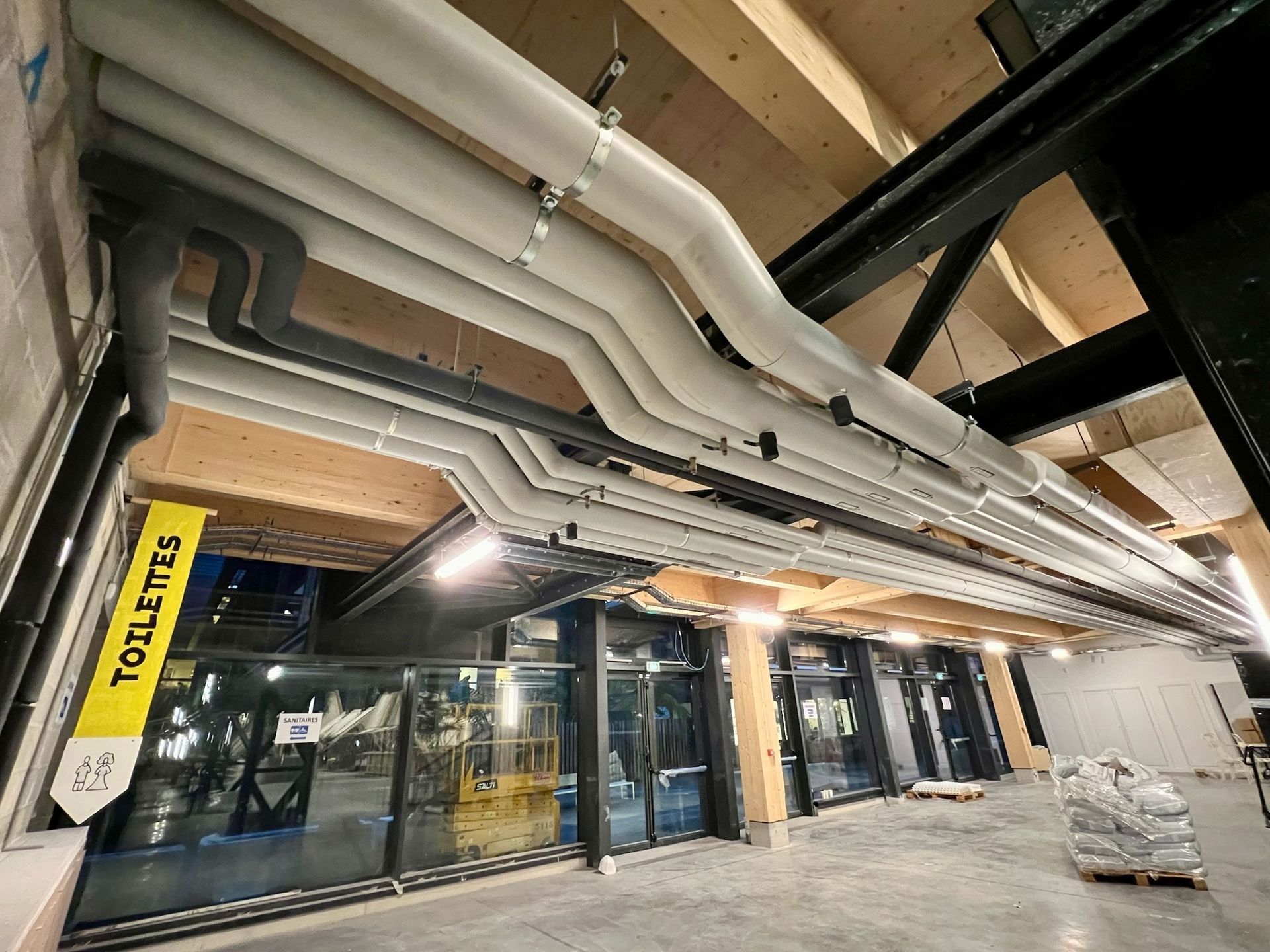 Des tuyaux et des conduits courent au plafond au-dessus de pièces vitrées. Un panneau « TOILETTES » est visible.