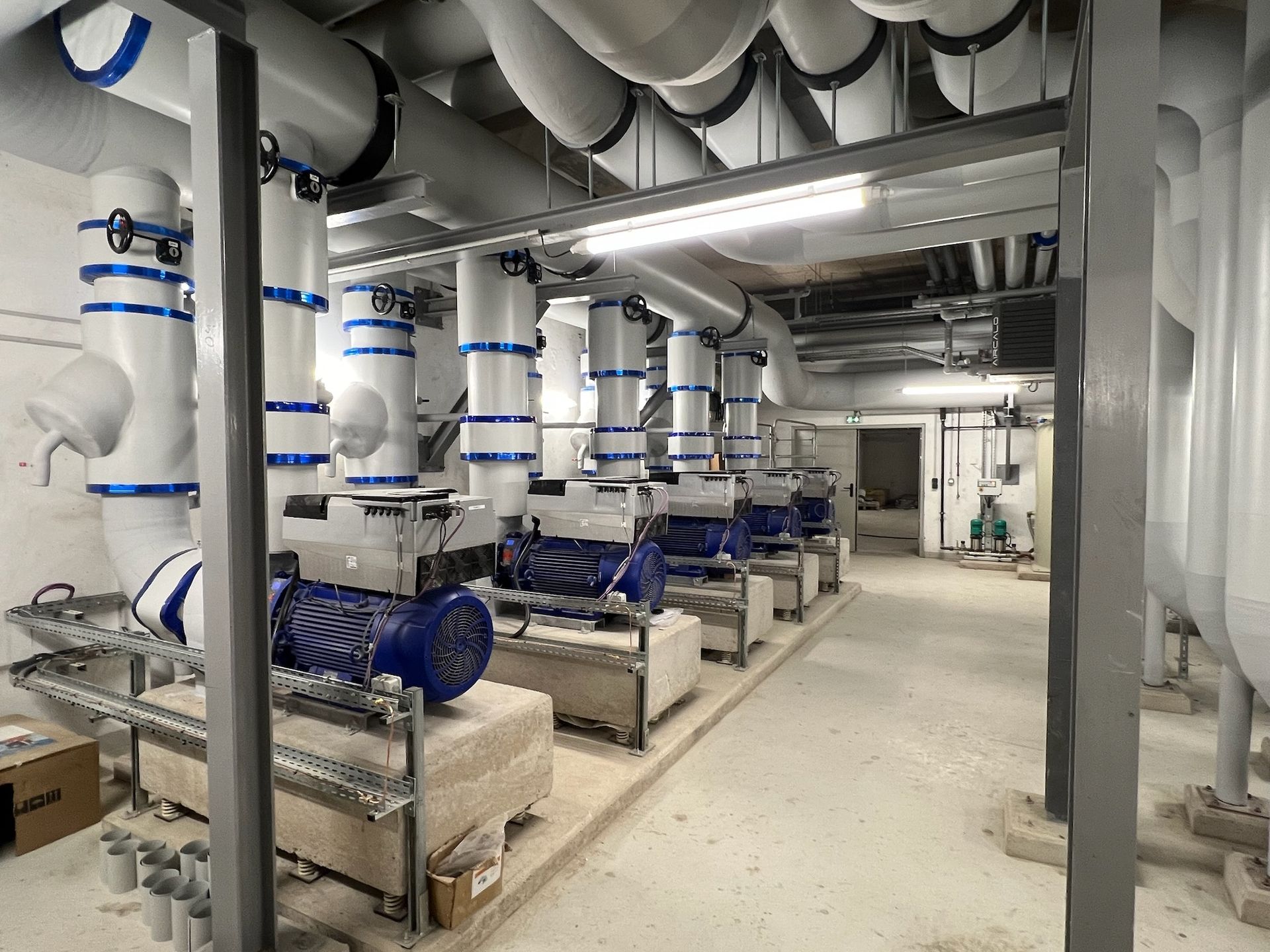 Rangée de pompes bleues dans une buanderie avec des tuyaux blancs et des poutres de support grises.