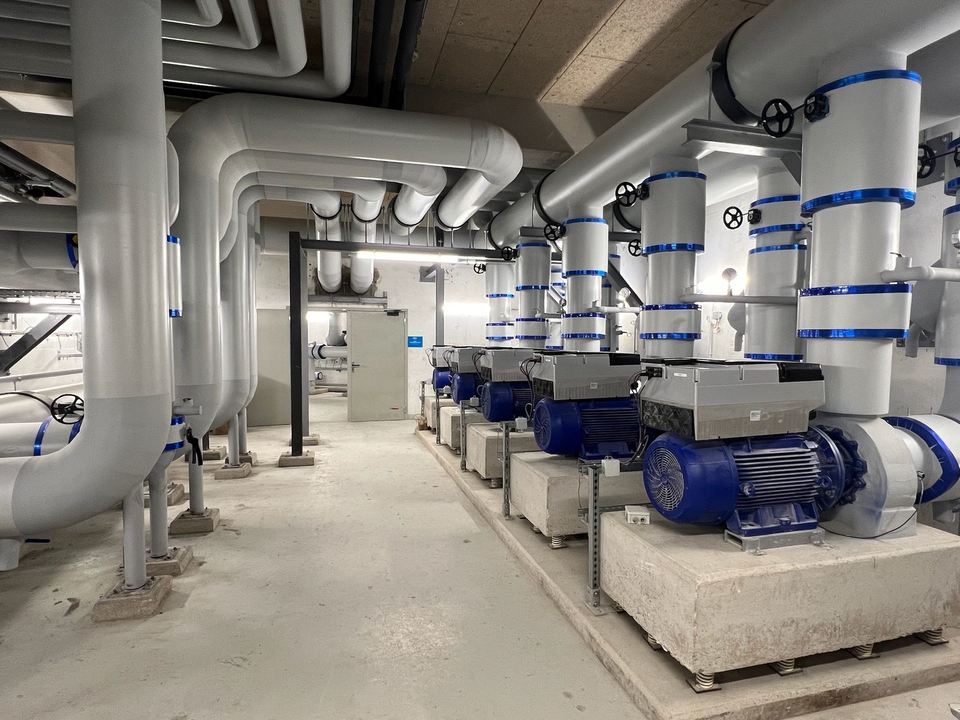 Installation industrielle avec rangées de pompes motorisées bleues et tuyaux blancs connectés, à l'intérieur.