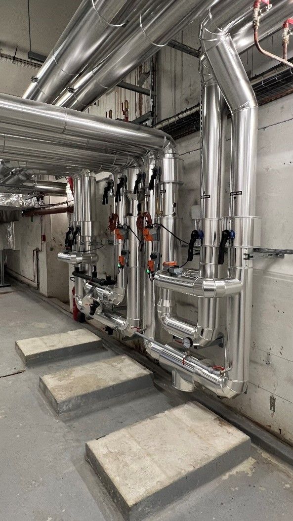 Tuyauteries dans une buanderie, avec isolation argentée. Sol en béton, murs gris.