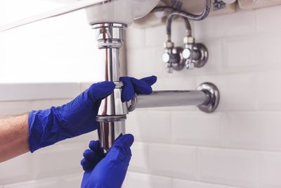 Des mains gantées de bleu réparent un tuyau d'évacuation sous un lavabo.
