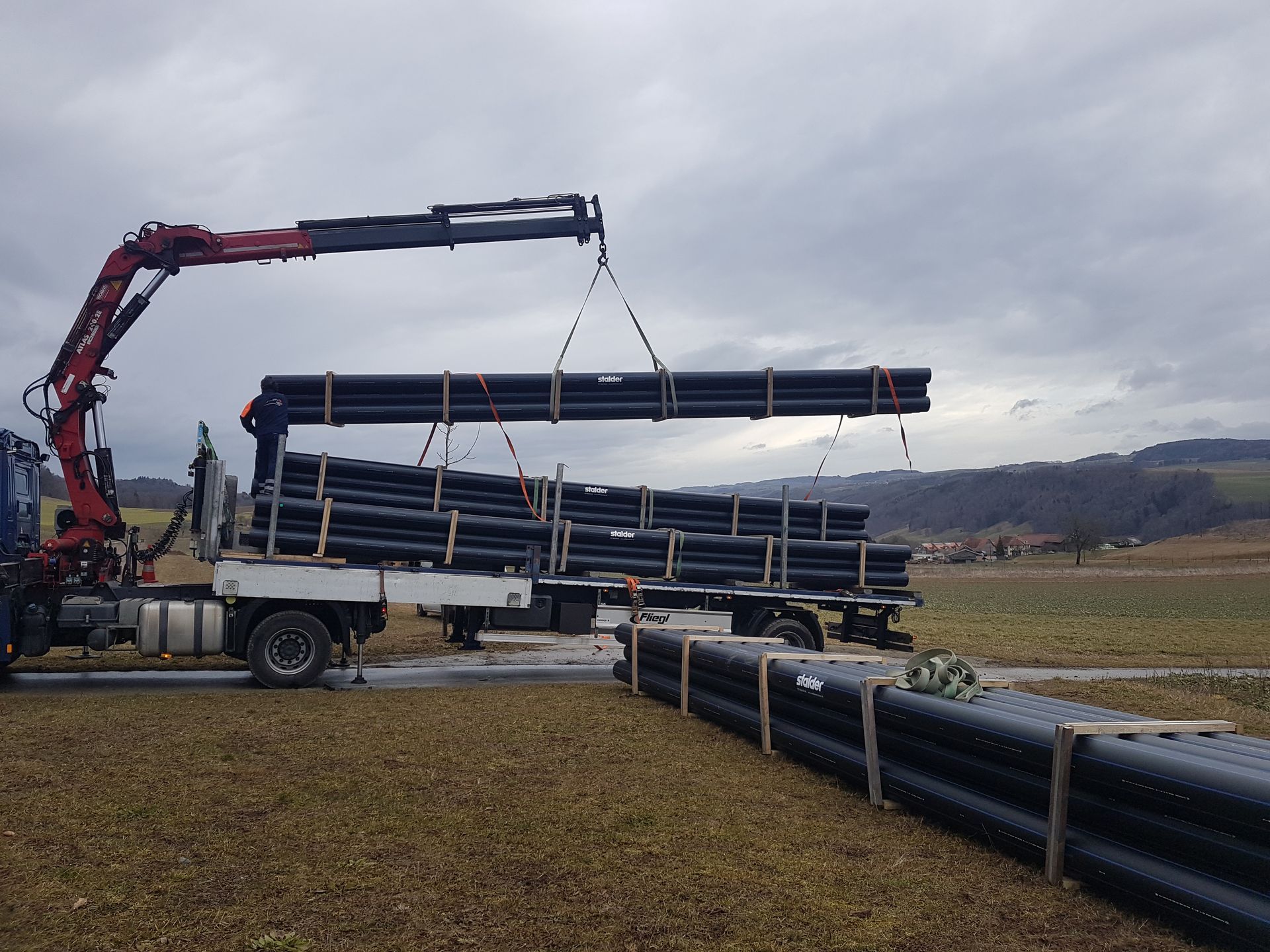 Une grue charge de longs tuyaux noirs sur un camion plateau dans un champ ouvert sous un ciel nuageux.