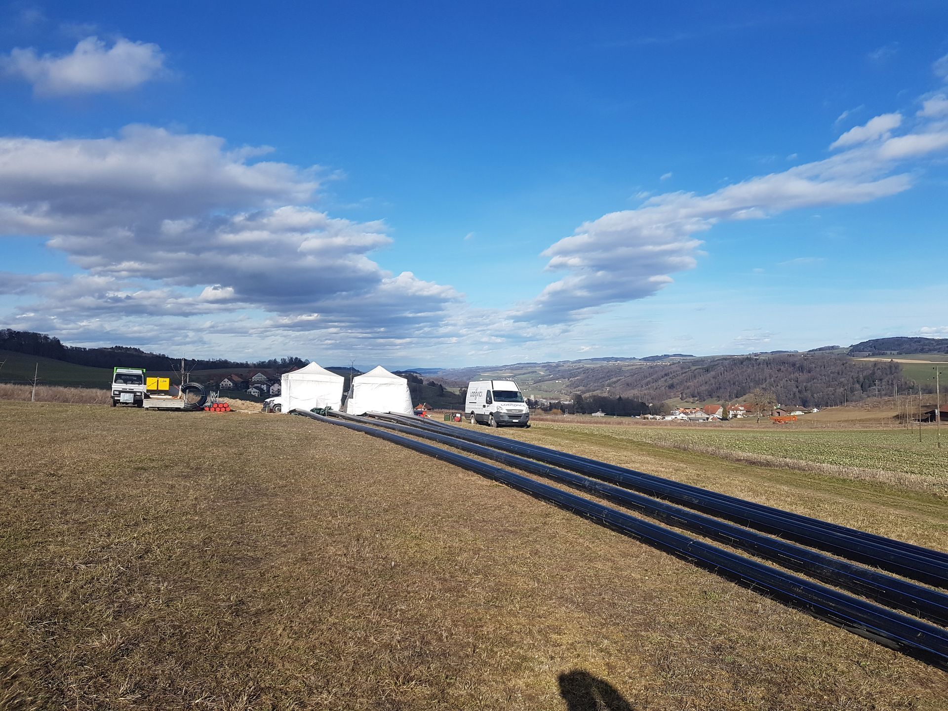 Champ rural avec de longs tuyaux noirs, des tentes blanches et des véhicules sous un ciel bleu nuageux.