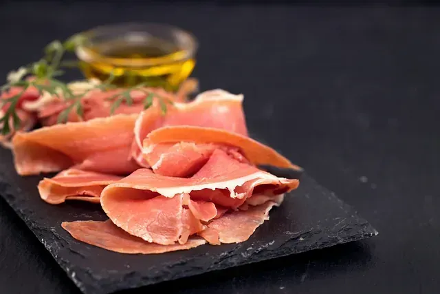 Jamón cortado en lonchas sobre una tabla de cortar de pizarra junto a un vaso de aceite de oliva.