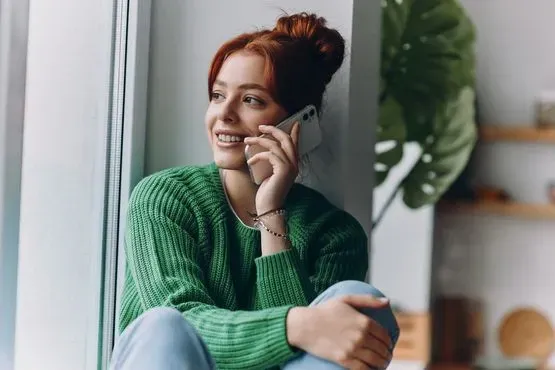Una persona con un suéter verde sonríe mientras habla por teléfono junto a una ventana.