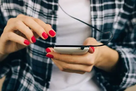 Manos con las uñas pintadas de rojo usando un teléfono inteligente con auriculares puestos.