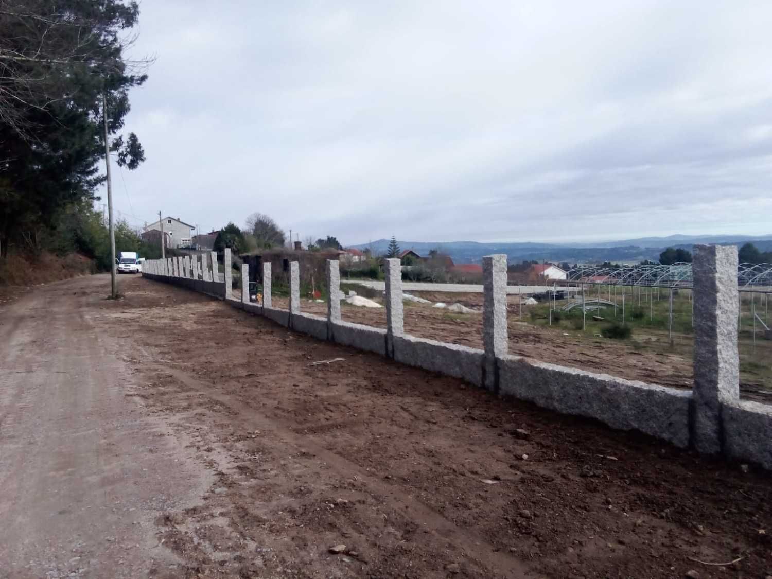 Muro de piedra a lo largo de un camino de tierra; cielo nublado al fondo, campos y casas visibles en la distancia.