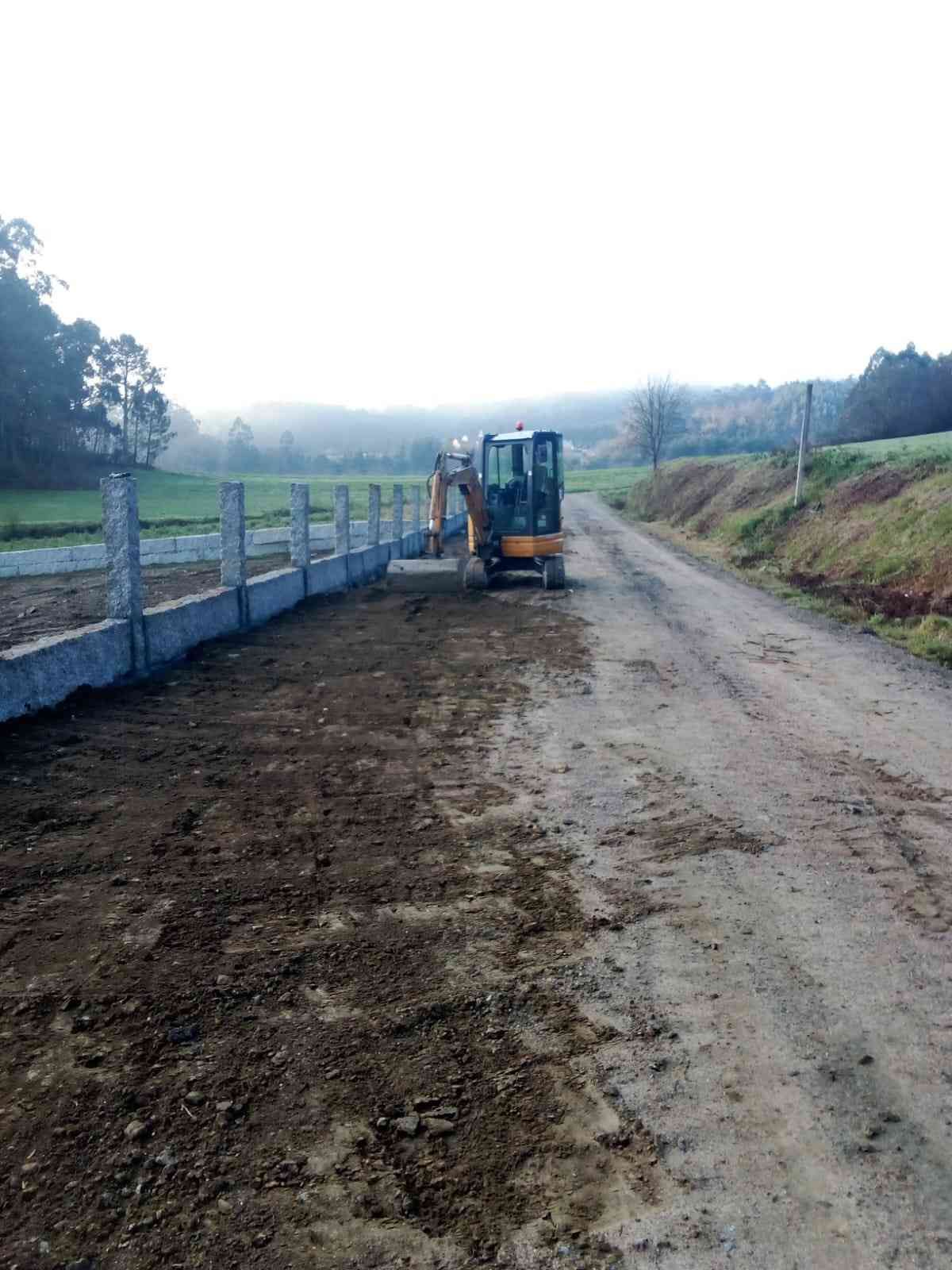 Miniexcavadora estacionada cerca de una valla de hormigón y un camino de tierra en un entorno rural. Cielo nublado.