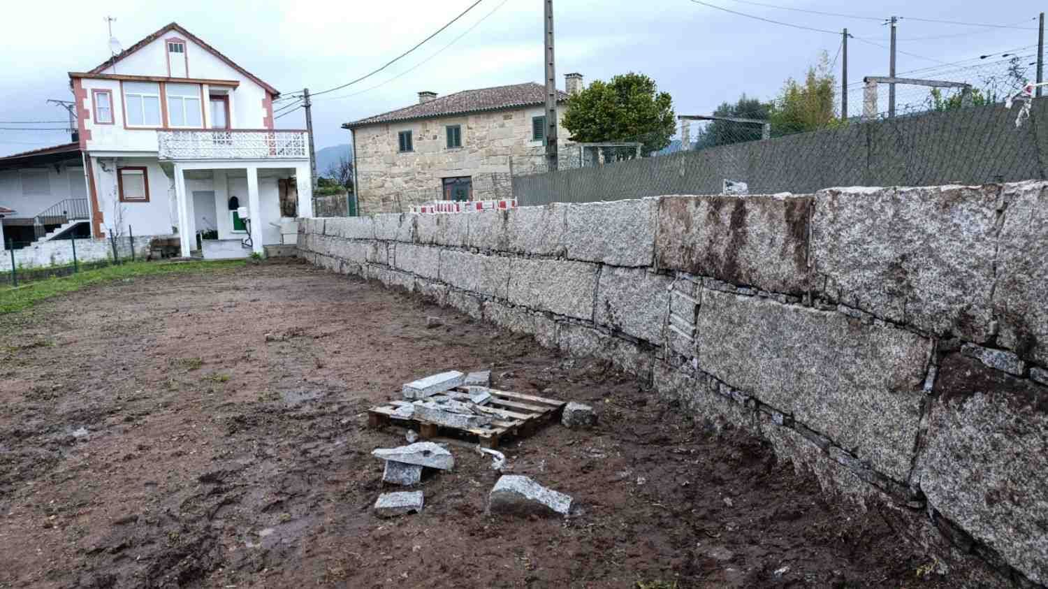 Muro de piedra de granito curvado frente a una casa, con escombros en la base, bajo un cielo nublado.