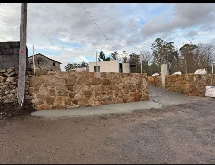 Muro de piedra y rampa de hormigón que conduce a un edificio blanco; entorno rural, cielo nublado.